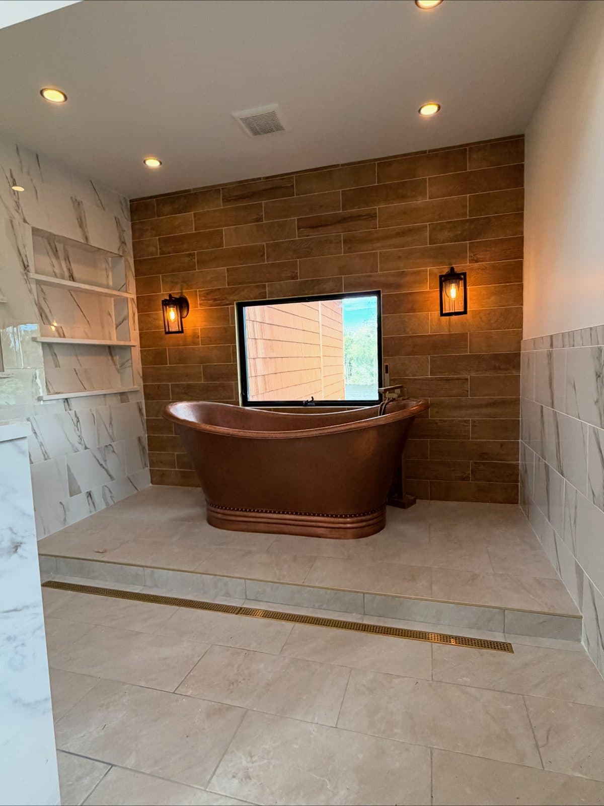 A remodeled bathroom lit by recessed ceiling lights and two wall sconces either side of the window over a copper tub