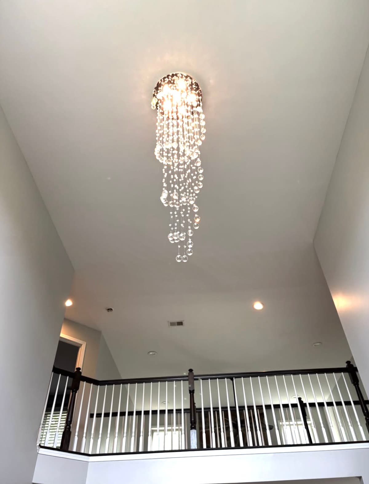 A spiral crystal chandelier lit high on the ceiling of a two-story foyer above a dark wood stair railing
