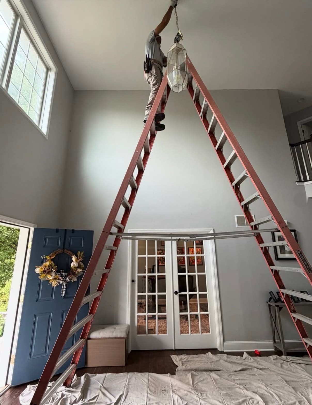 An electrician at the top of a tall extension ladder hanging a glass lantern fixture from a two-story foyer ceiling, drop cloths on the floor